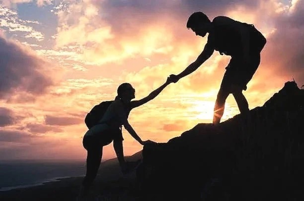 Two hikers helping each other up a sunset ridge